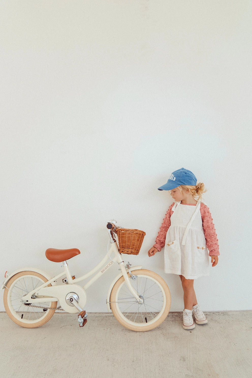 Preschool child standing next to a white bicycle with a basket against a plain white wall.