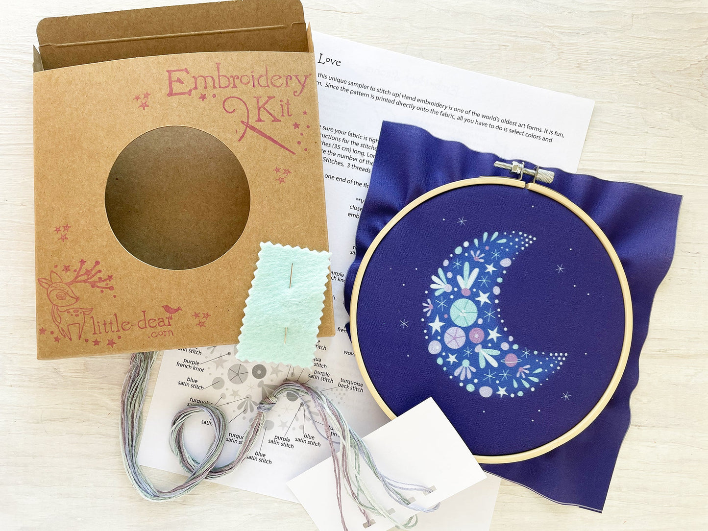 Embroidery kit with a completed design on a blue fabric hoop, surrounded by packaging and instructions.