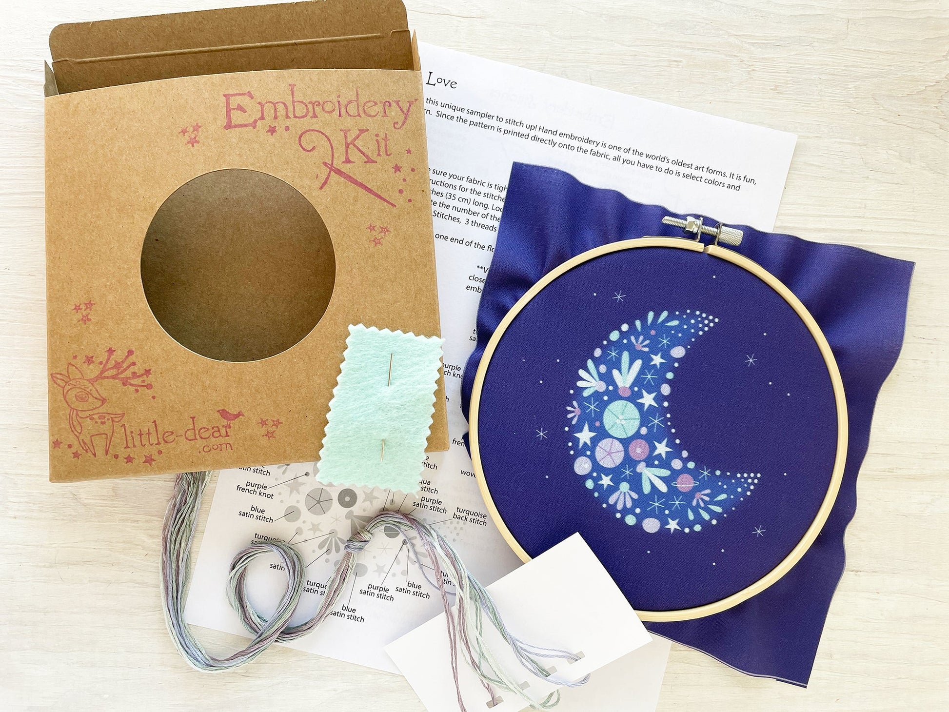 Embroidery kit with a completed design on a blue fabric hoop, surrounded by packaging and instructions.