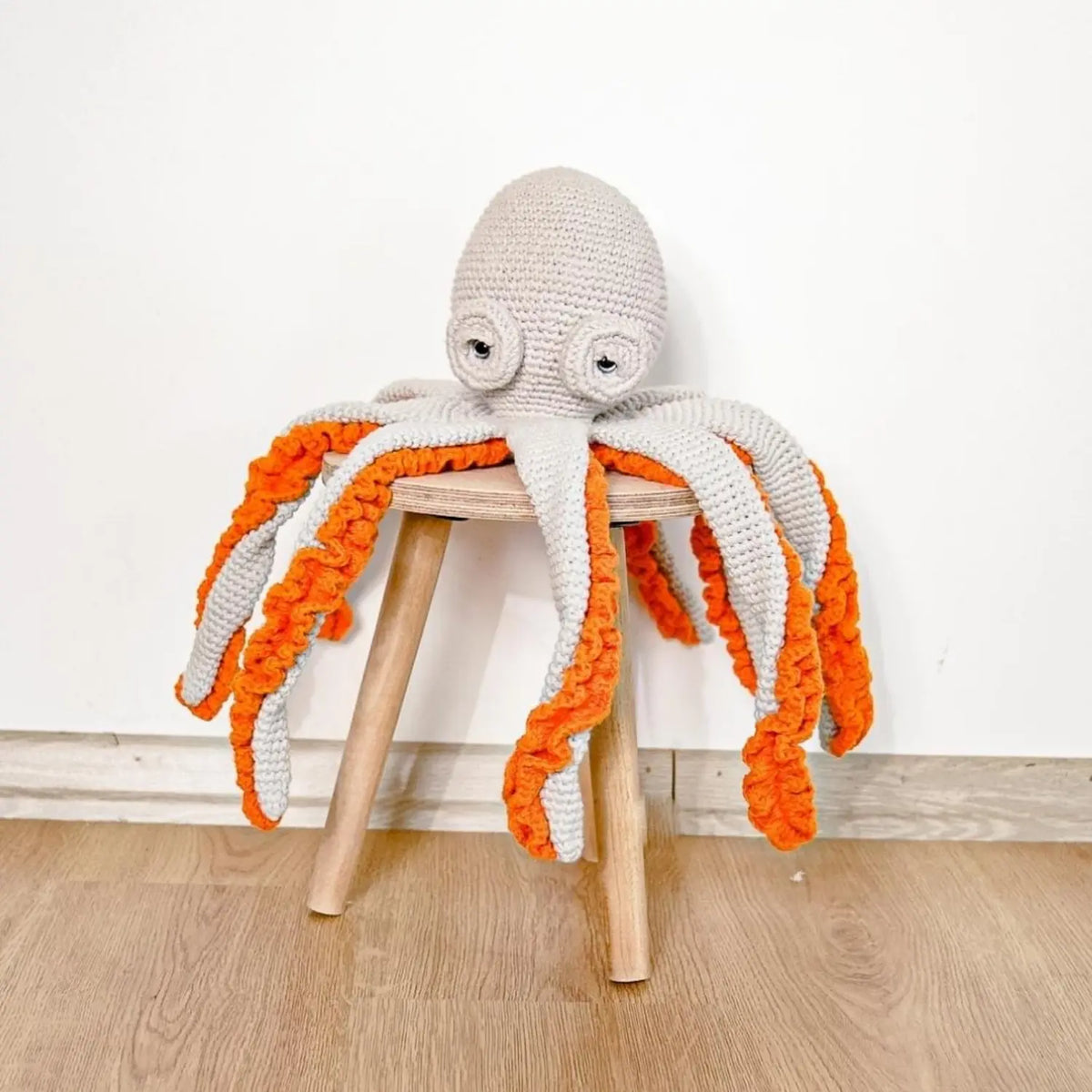 Light gray crochet octopus soft toy sitting on a small wooden stool, its long orange-ruffled arms draping to the floor