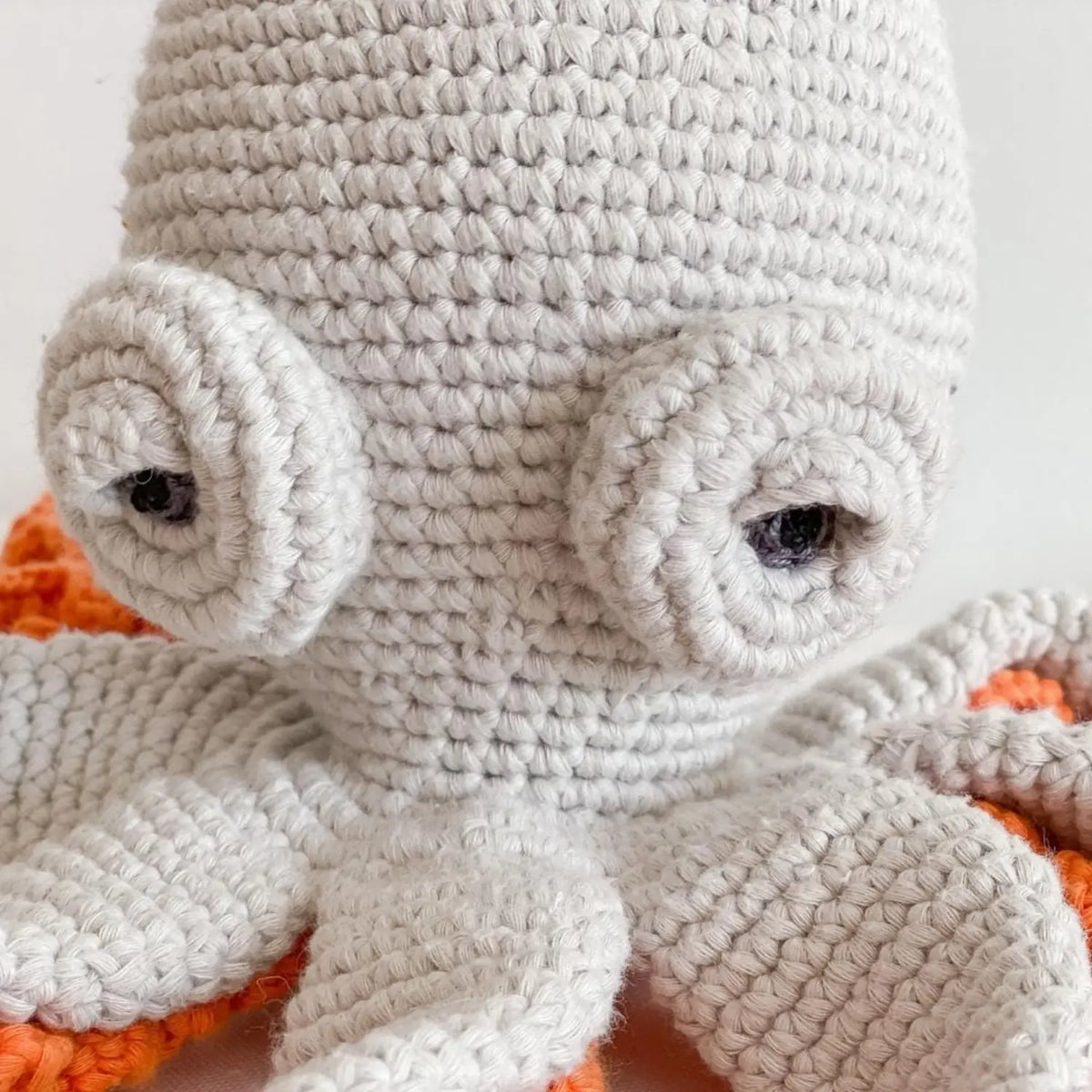 Close-up of the light gray crochet octopus's spiral eyes and hand-crocheted organic cotton stitches, with orange ruffles behind
