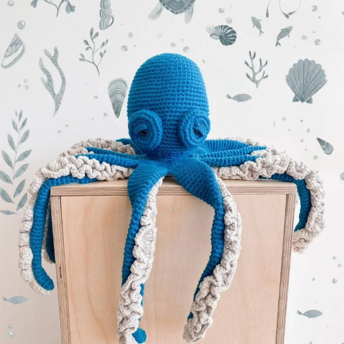 Lagoon teal crochet octopus soft toy draped over a wooden cube, its ruffled arms hanging down against an ocean-print wall