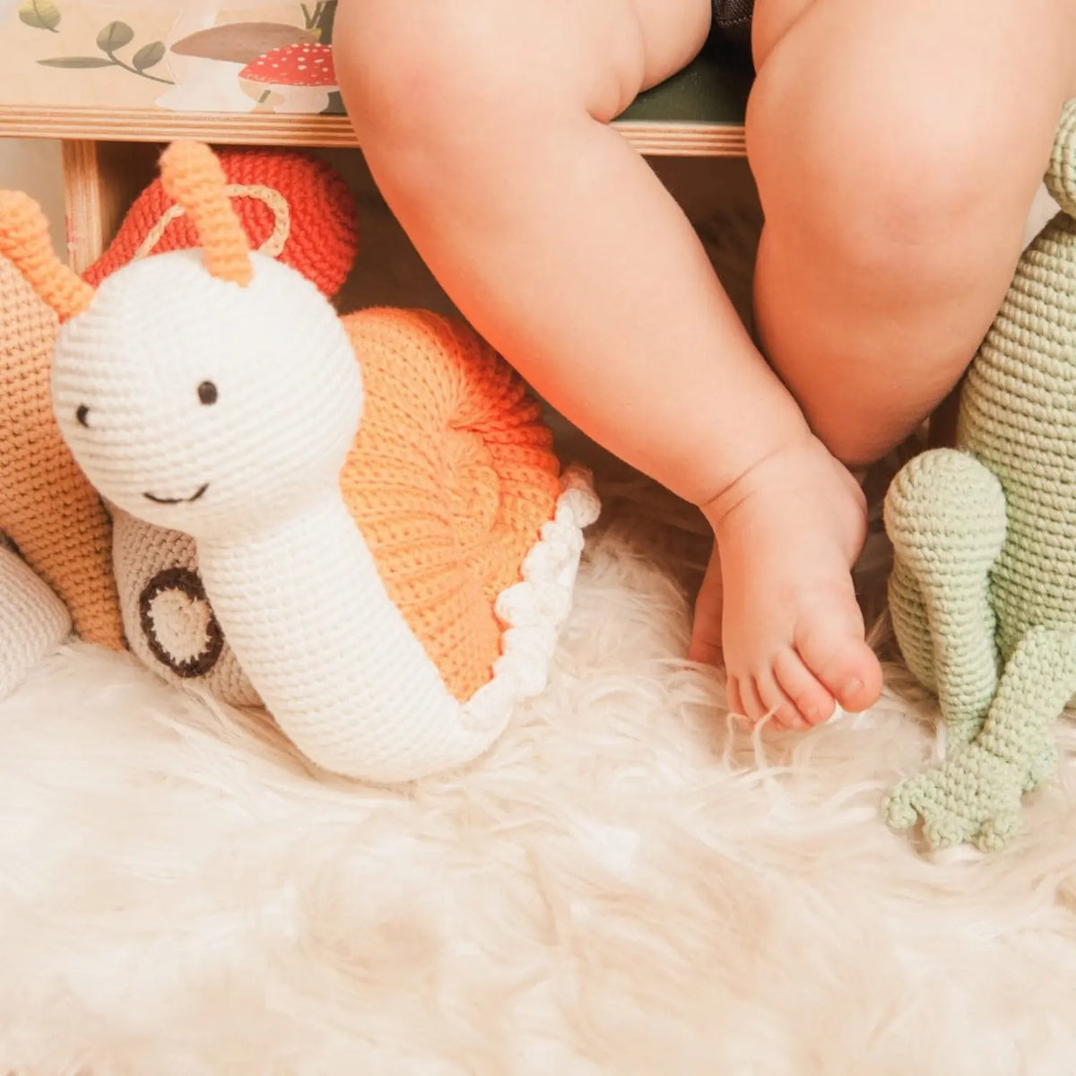 Crochet snail soft toy on a sheepskin rug beside a baby's legs and feet, with other crocheted animals nearby