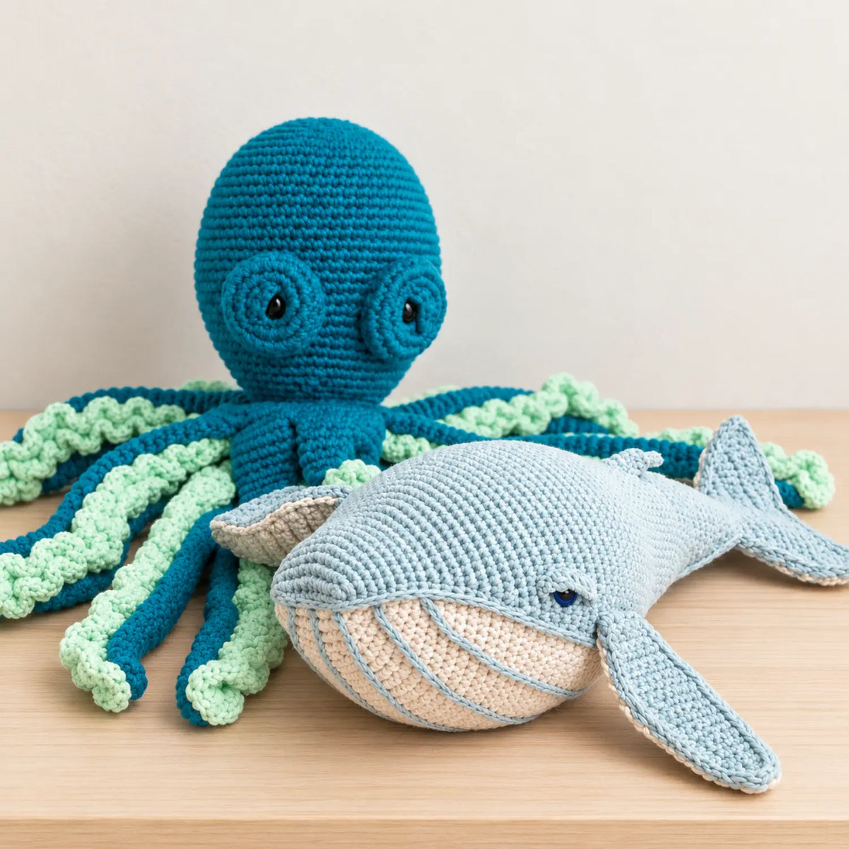 Misty blue crochet whale soft toy in front of a teal and mint crochet octopus with curly tentacles on a wooden table