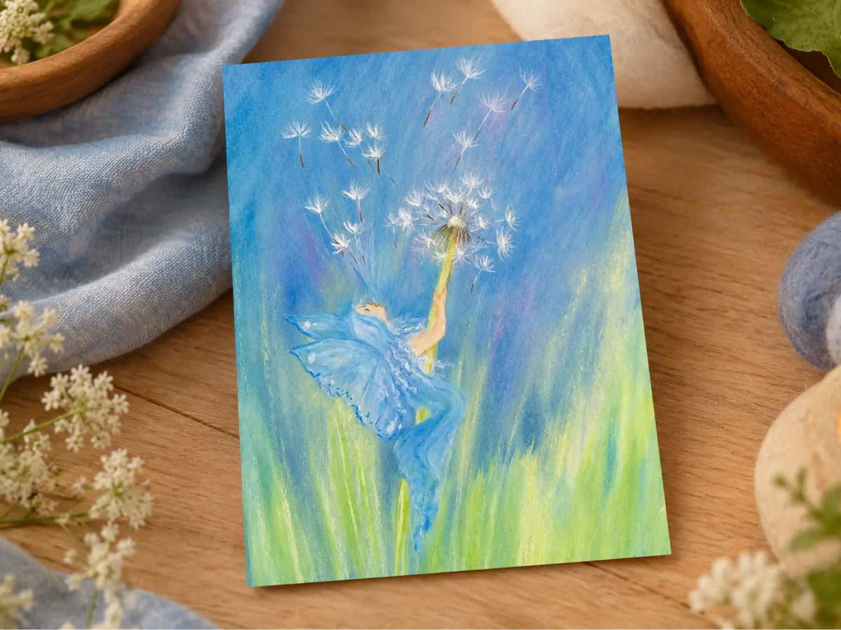 Marjan van Zeyl Dandelion Fairy postcard displayed on a wooden table with soft blue linen, white flowers, and natural seasonal decorations.