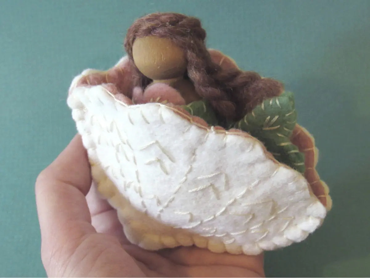 Hand holding a Waldorf mermaid peg doll in a felt seashell with brown hair against a teal background