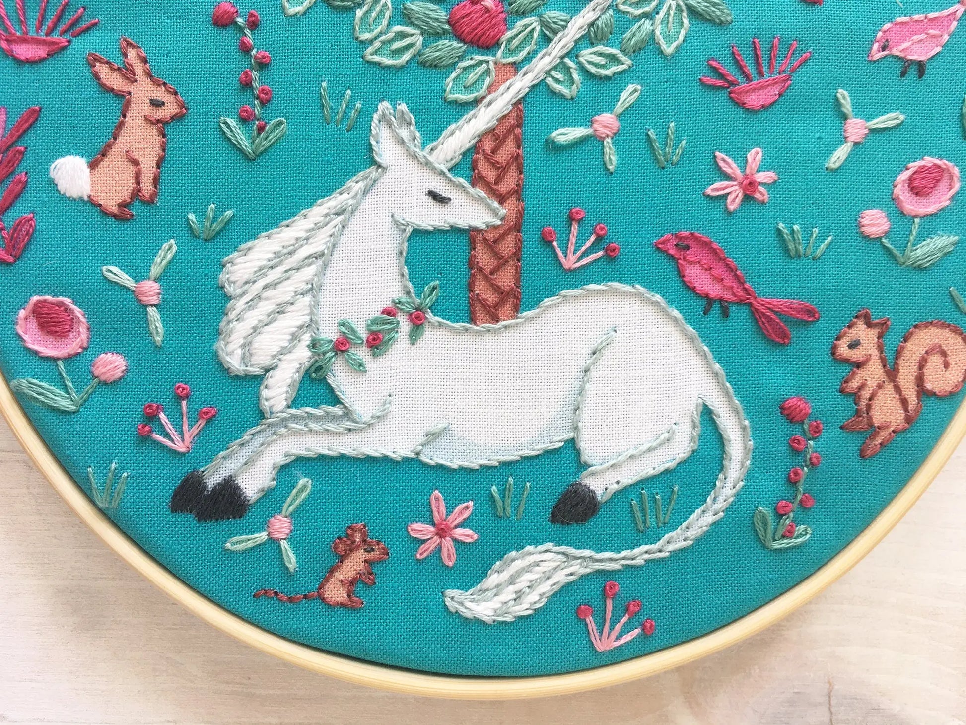 Embroidery hoop with a unicorn design surrounded by flowers and animals on a teal background