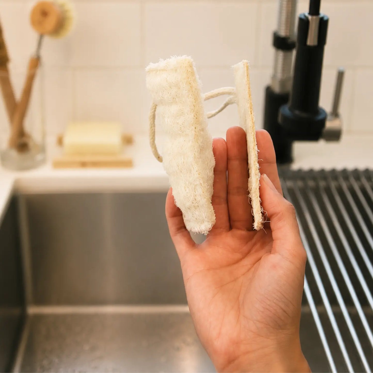 Hand holding a double-layer loofah dish sponge beside a side view to show its thickness and plant fiber construction.