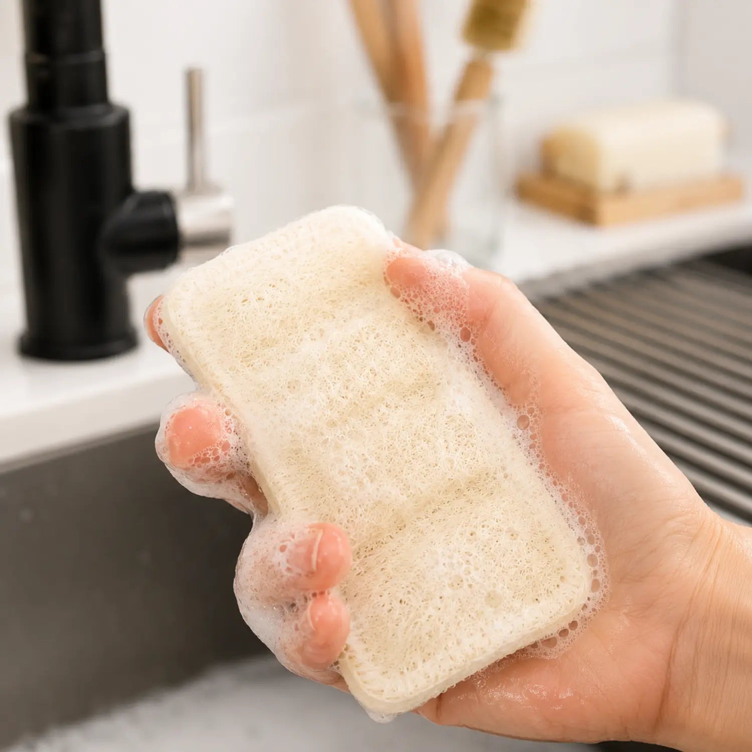 Wet double-layer loofah dish sponge with soap suds held in hand during kitchen dishwashing.