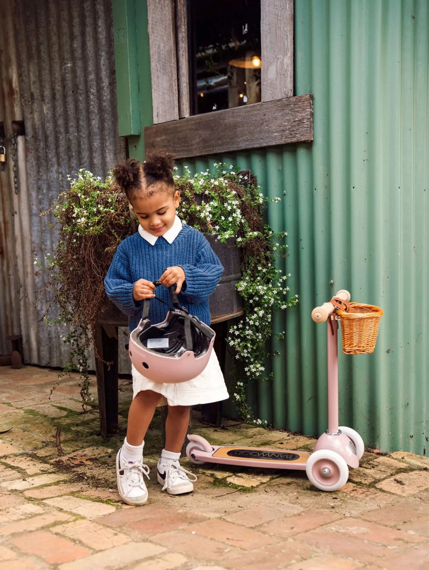 Child with a pink scooter and helmet in front of a rustic building.
