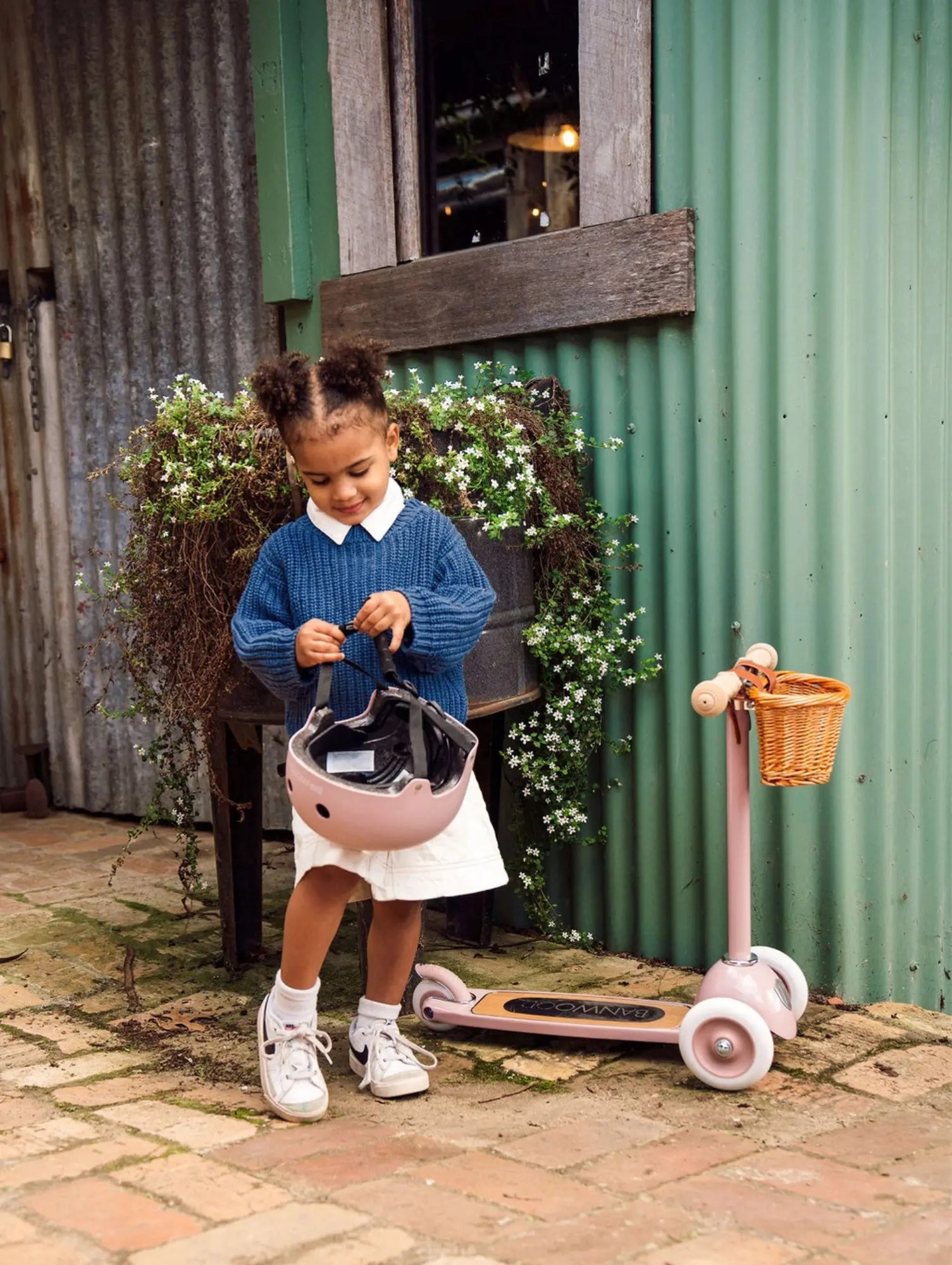 Child with a pink scooter and helmet in front of a rustic building.