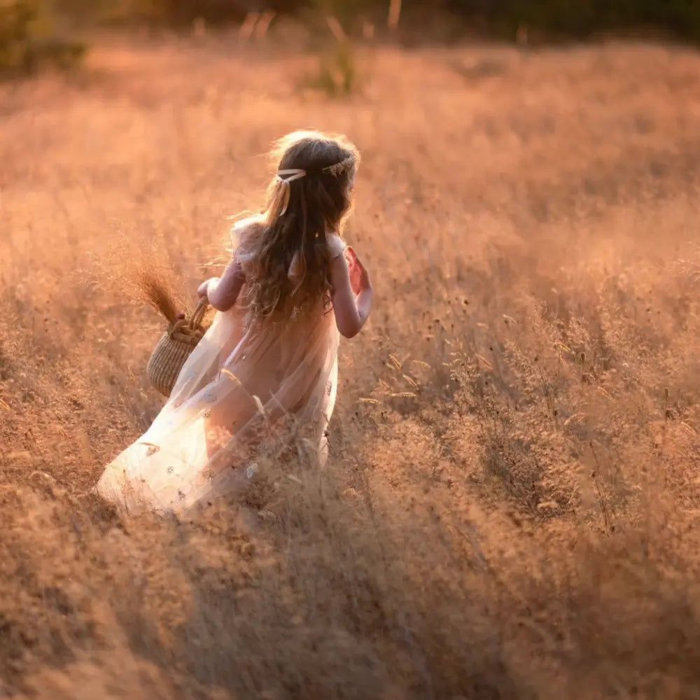 Young girl in a white dress walking through a field of tall grass at sunset.