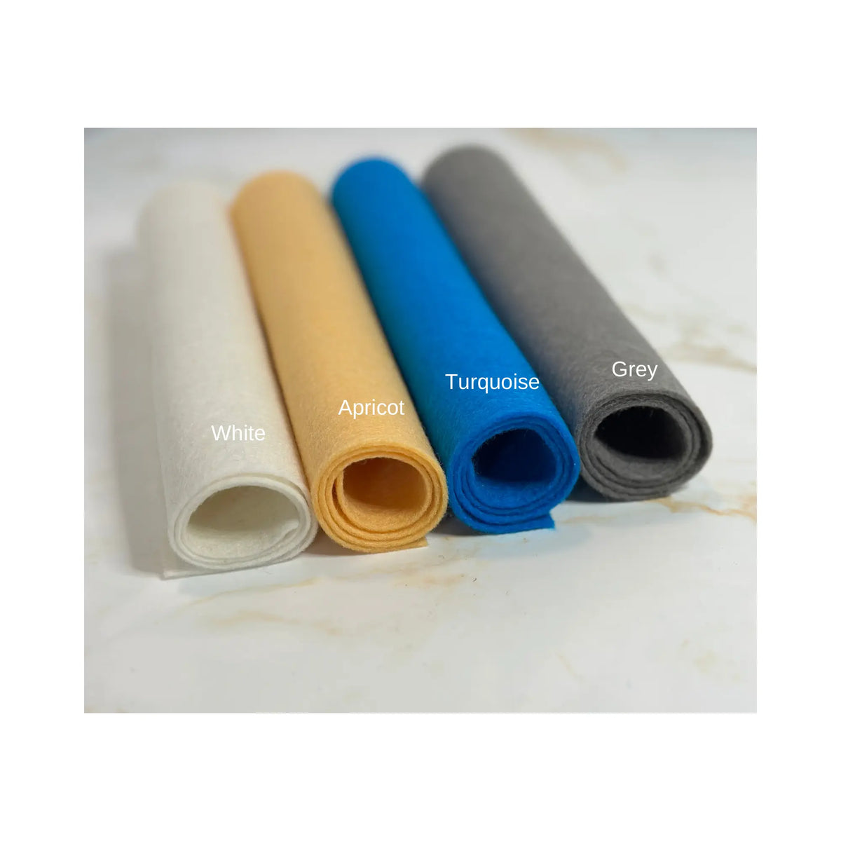 100% Wool Felt sheets rolled up on a marble surface in the colors white, apricot, turquoise, and grey