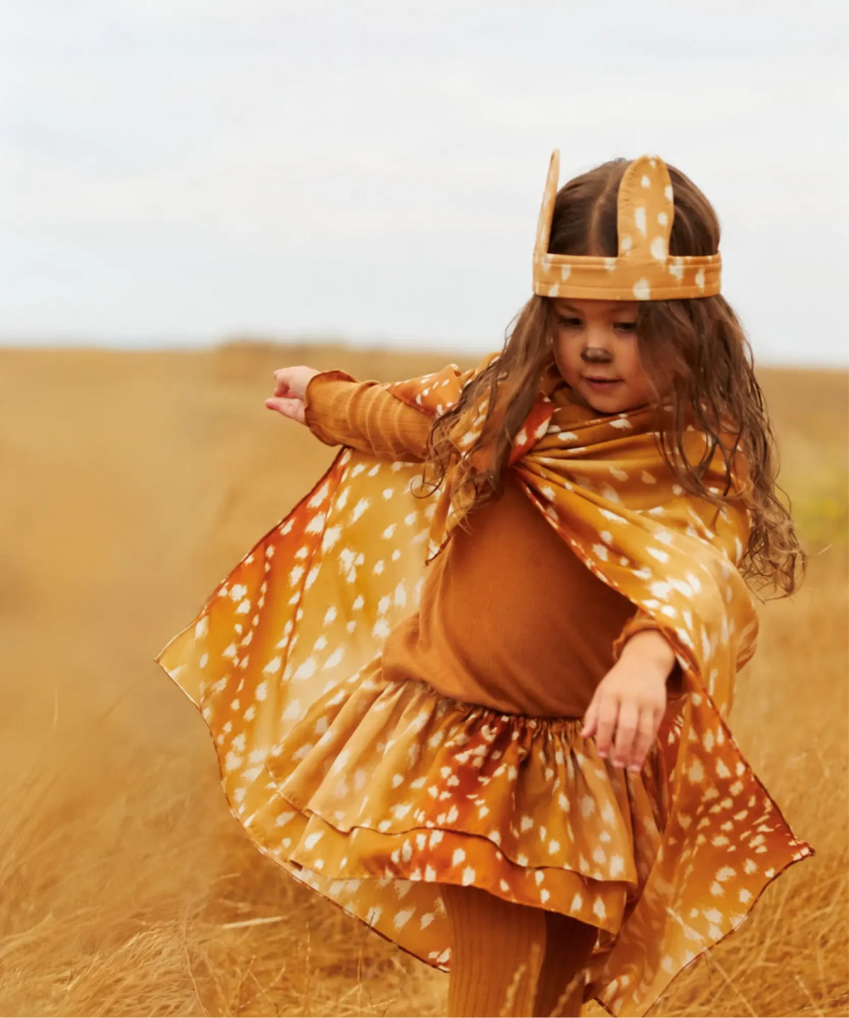 Child wearing fawn ears and a fawn-print Playsilk cape while playing in a field