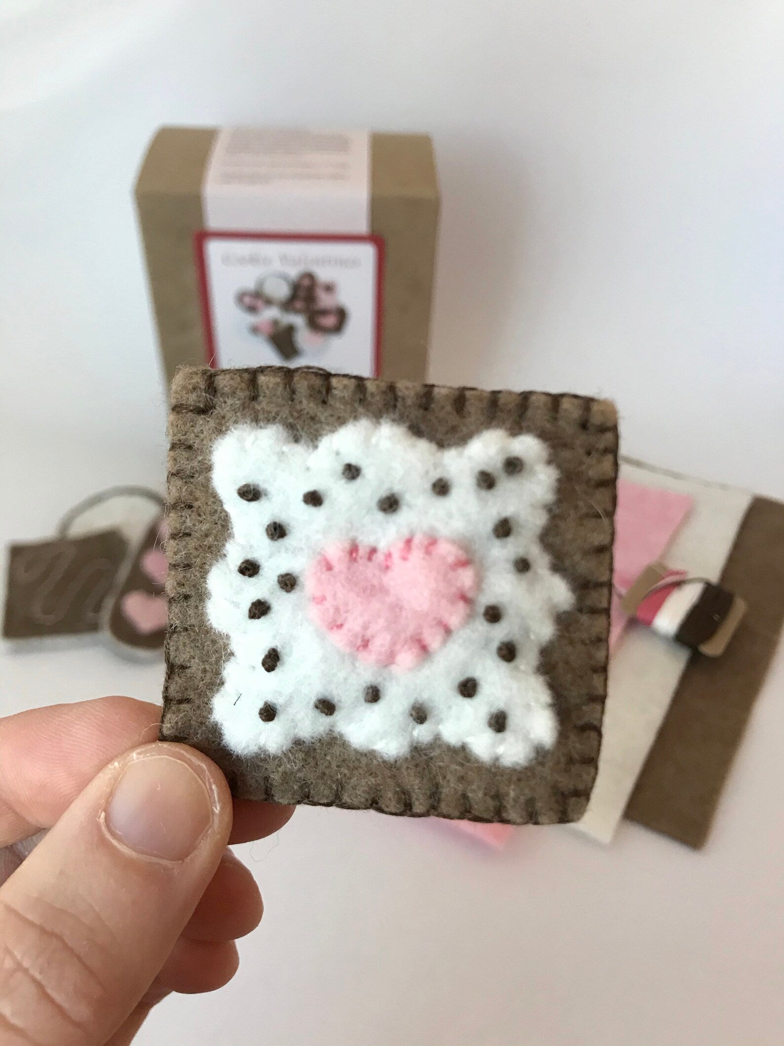 Handmade wool felt heart design on a square pretend play cookie held by a hand with a blurred background