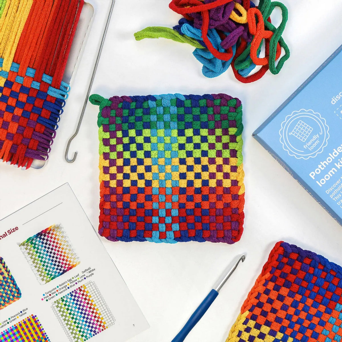 Finished rainbow checkerboard cotton potholder with a hanging loop, surrounded by loops, hooks and a pattern sheet