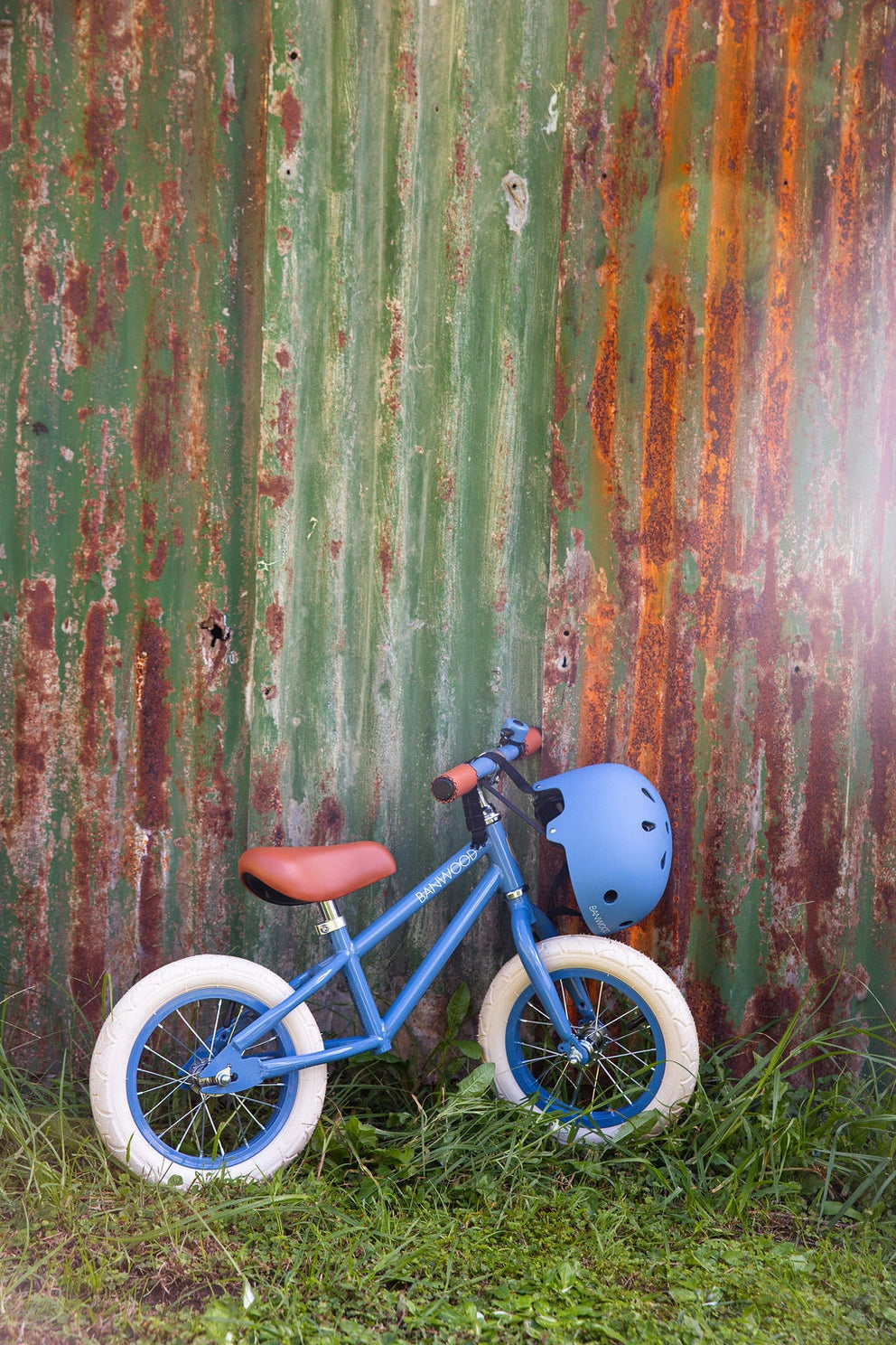 Blue balance bike with a helmet leaning against a rusty corrugated metal wall.