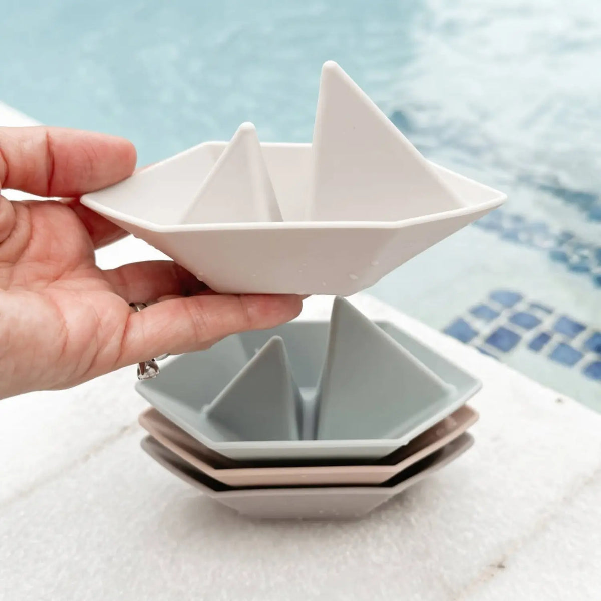 Hand holding a silicone floating boat bath toy above a stack of pastel boats near water.