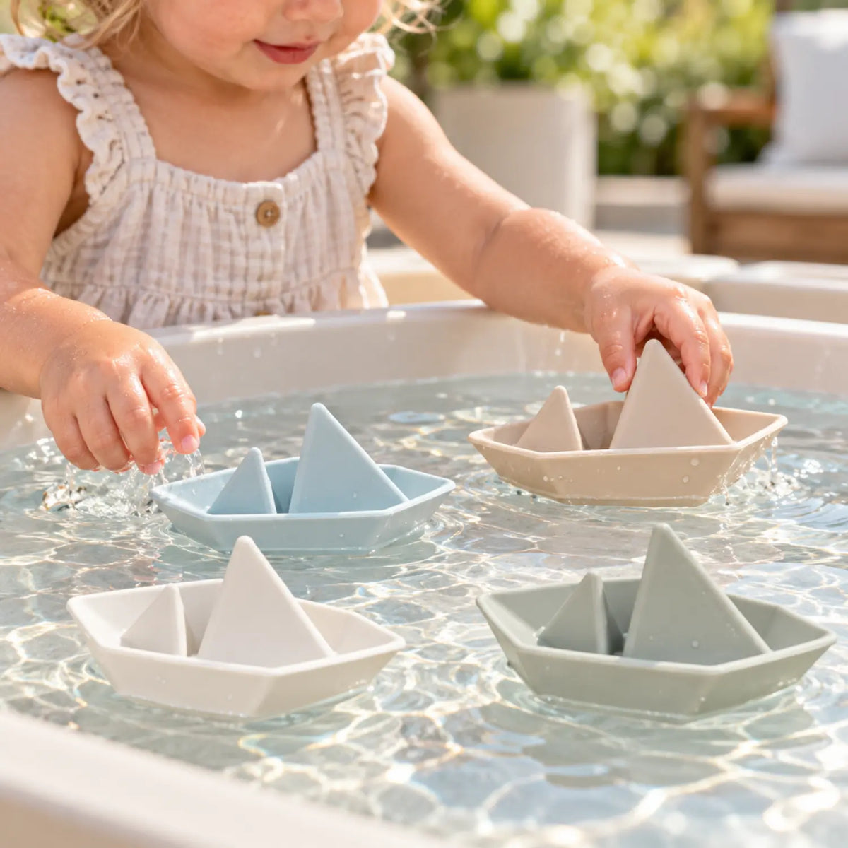 Toddler playing with four pastel silicone boat bath toys floating in a shallow outdoor water table.