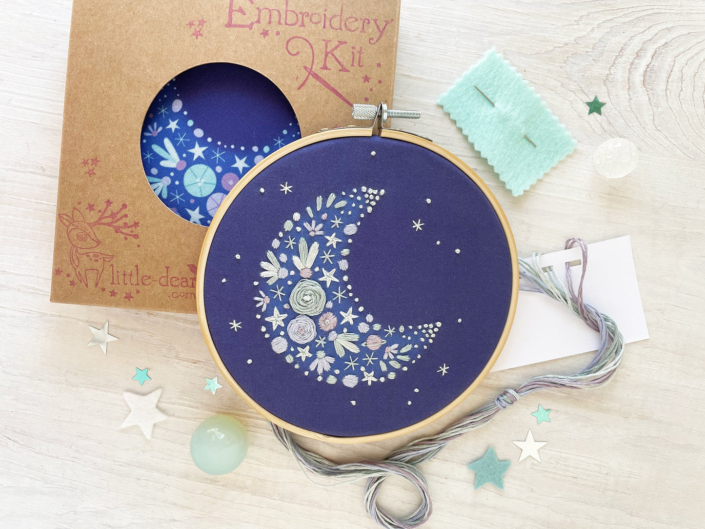 Embroidery kit with a completed design of a moon and stars on a blue fabric, surrounded by materials and packaging.