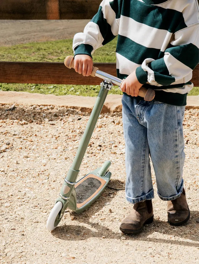 Child standing next to a scooter on a gravel path with grass in the background