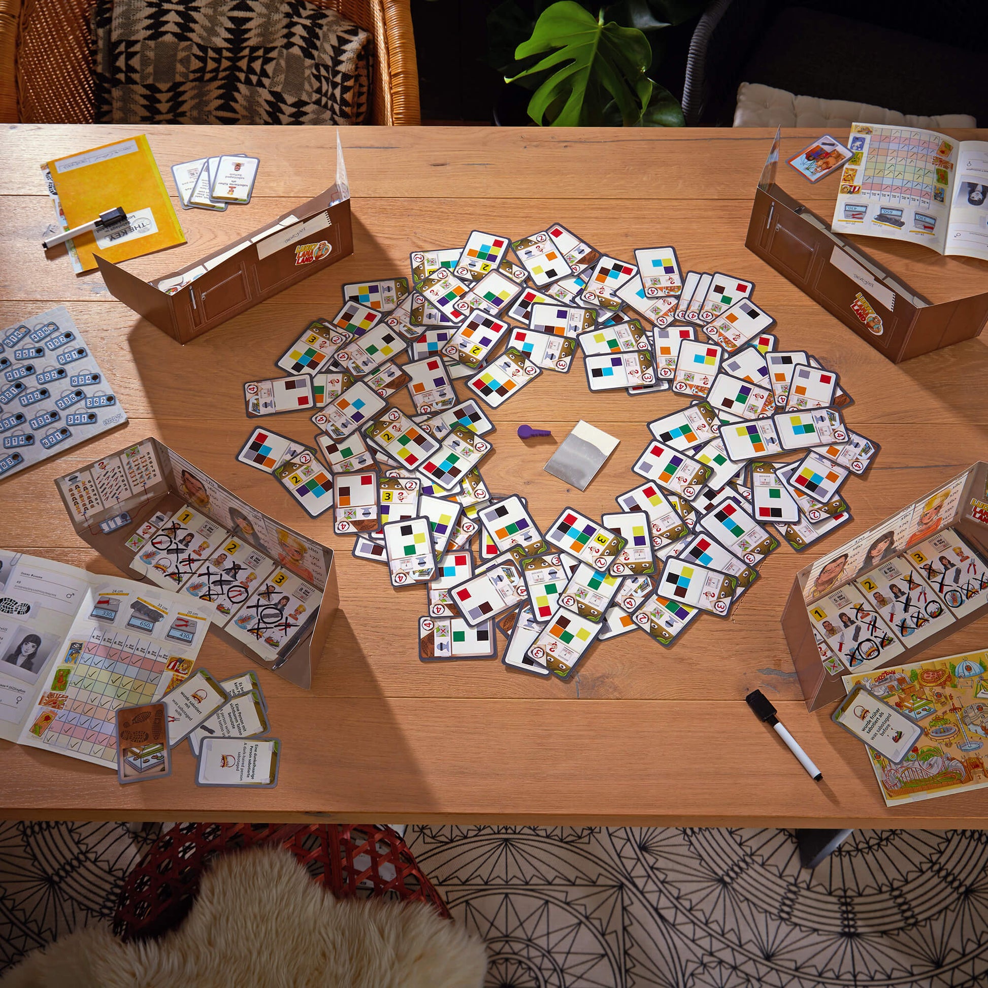 Scattered board game components on a wooden table with boxes and papers.