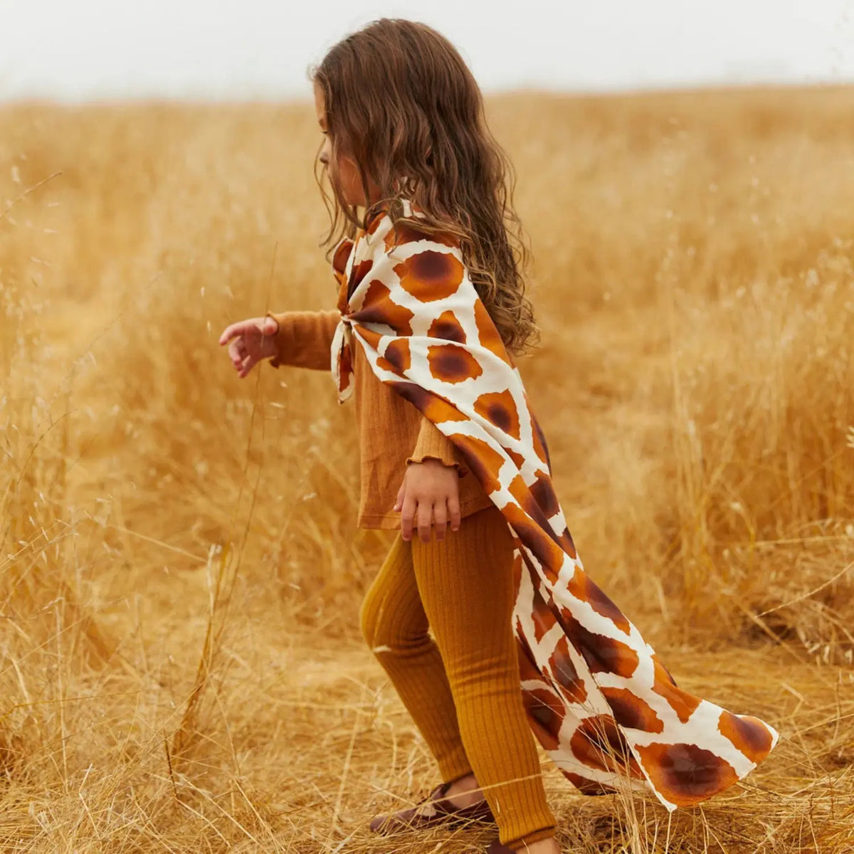 Child walking through a golden grass field wearing a giraffe Playsilk as a flowing cape