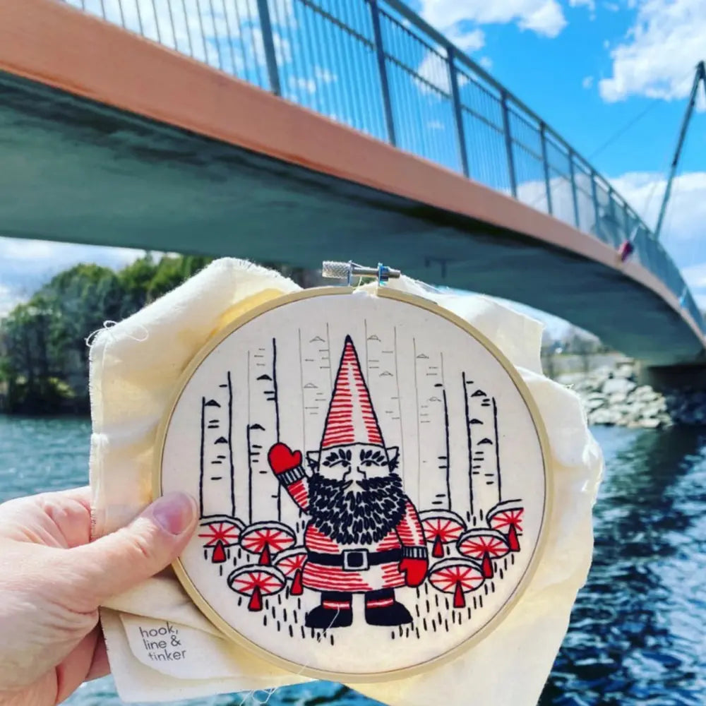 Gnome mushroom embroidery hoop held outdoors near a river and bridge
