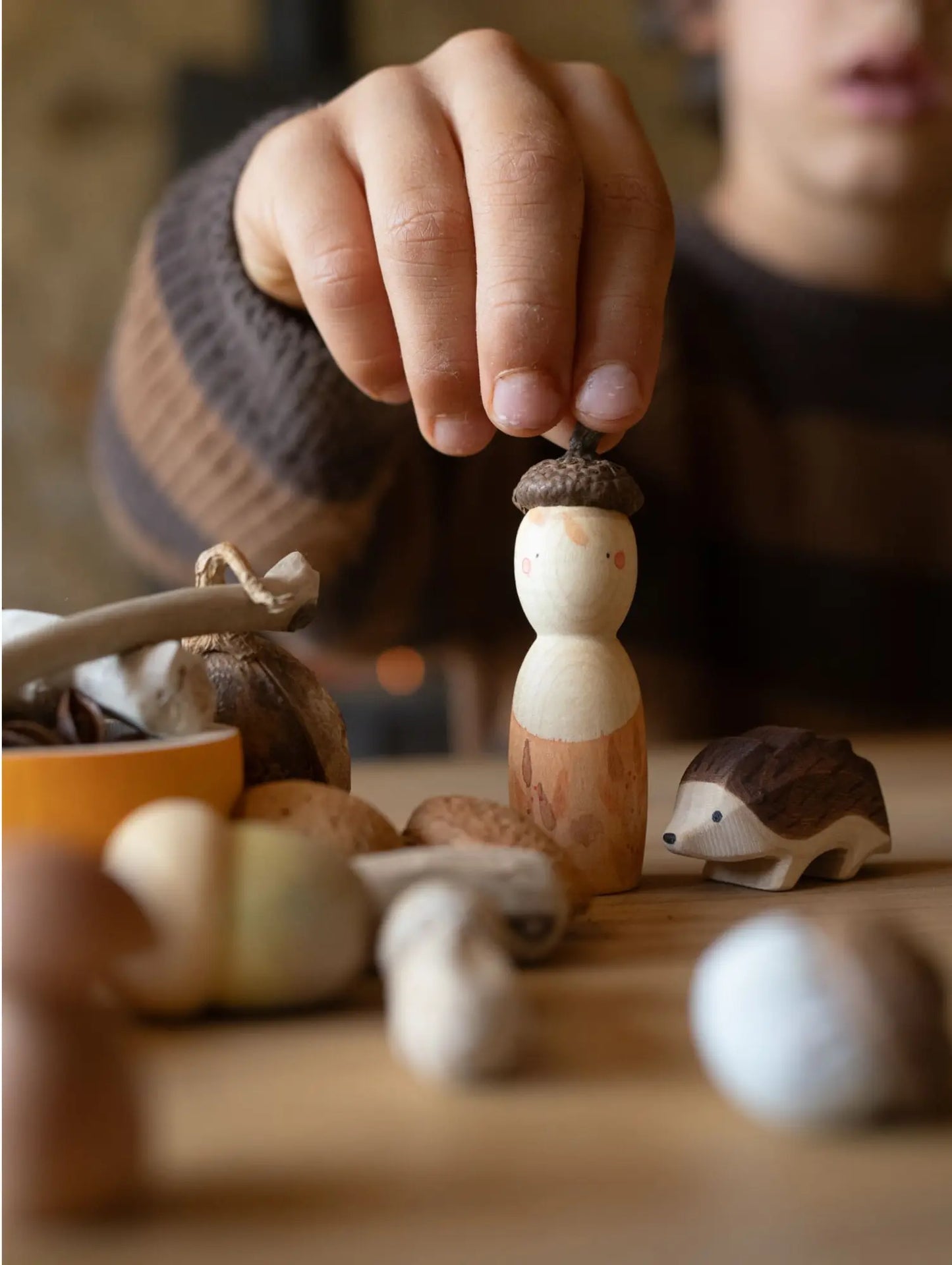 Child's hand holding a small acorn wooden toy figurine on a table with other decorative items.