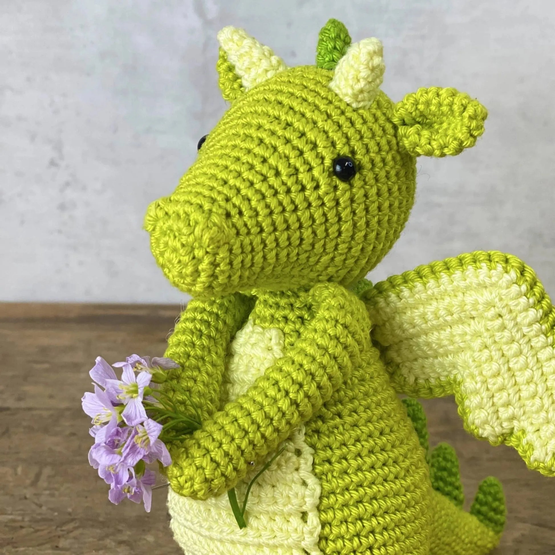 Full side view of green crocheted dragon soft toy with wings holding flowers
