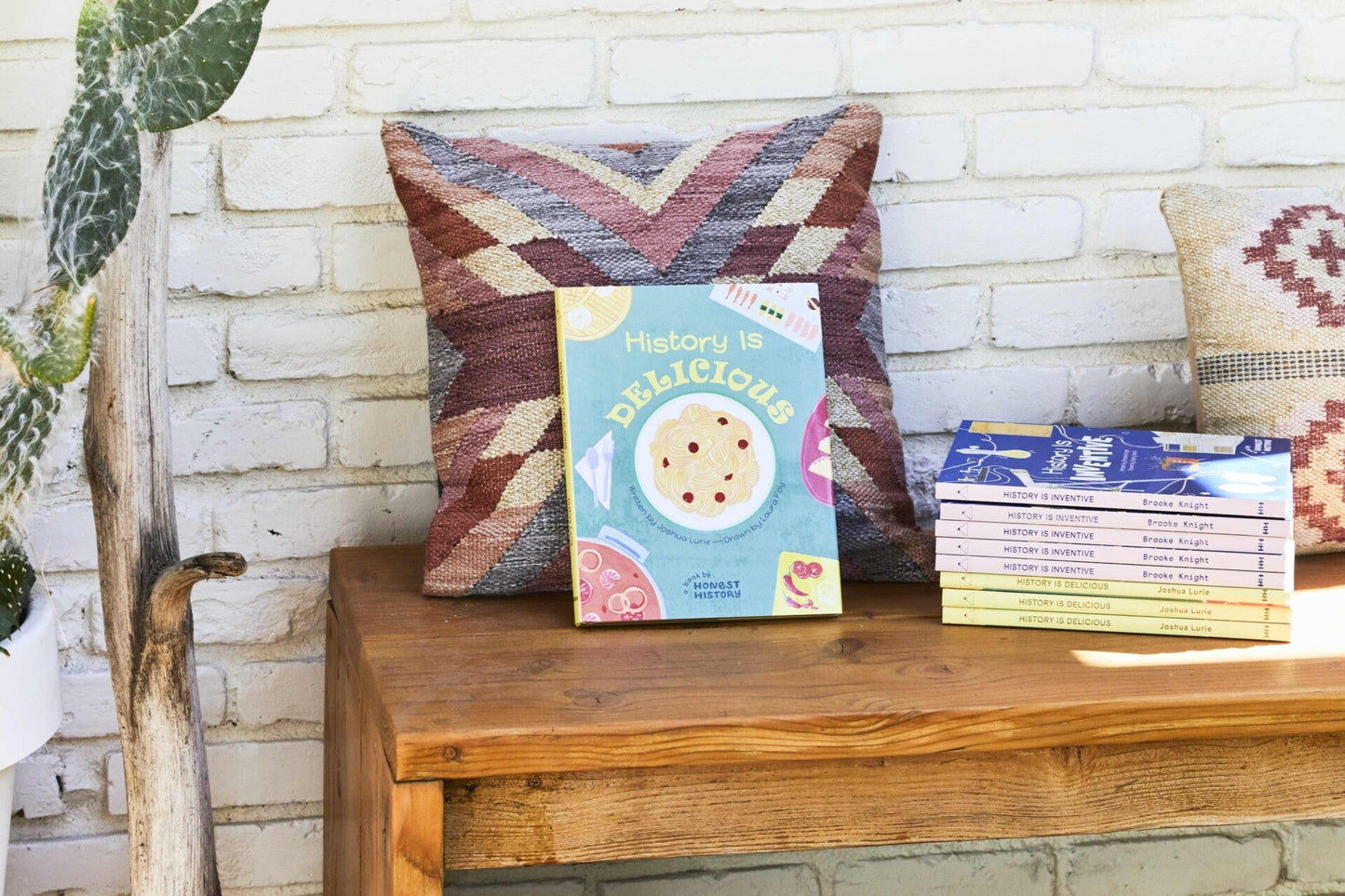 History Is Delicious children’s book displayed on a wooden bench with a pillow and a stack of other history books