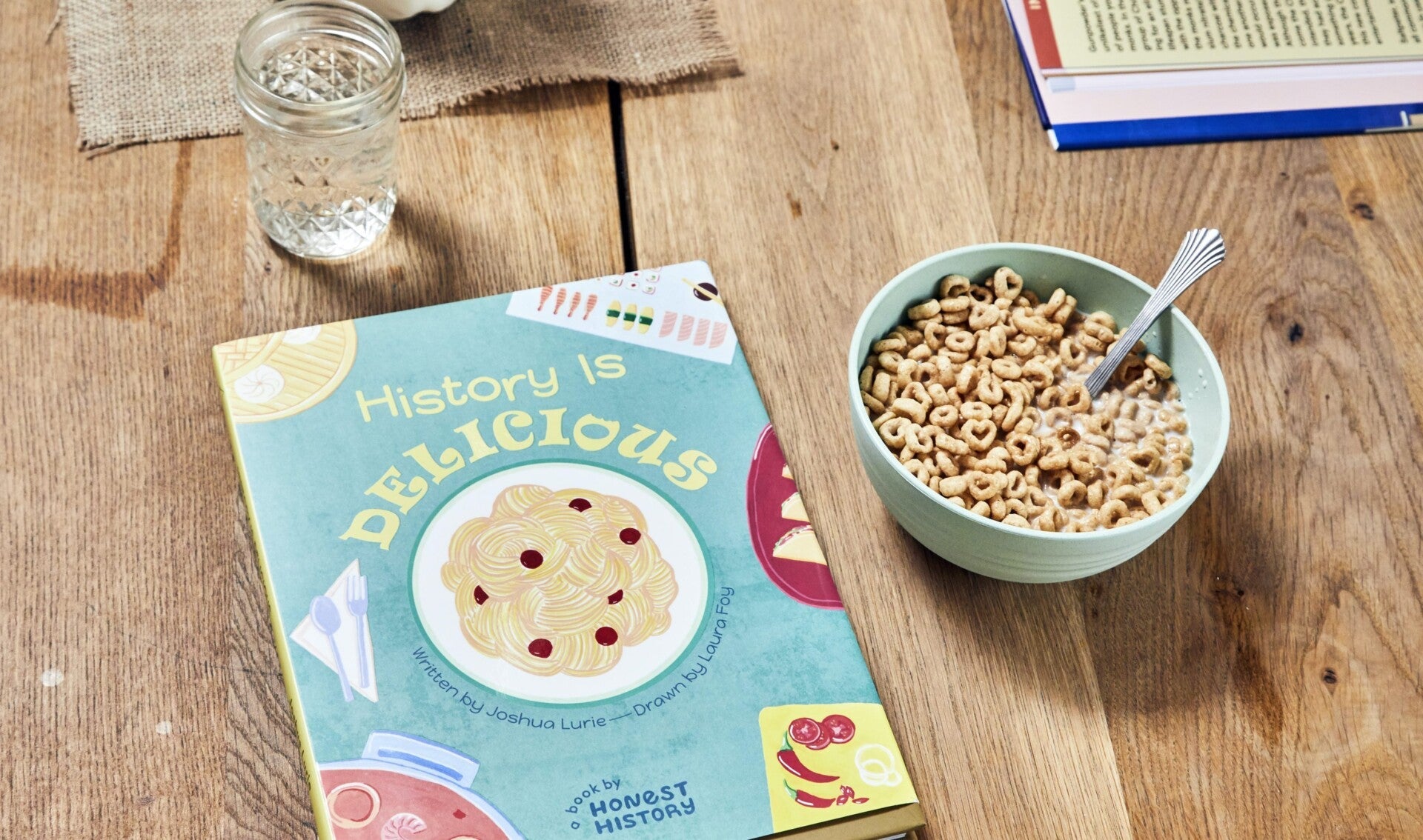 History Is Delicious children’s book displayed on a wooden table next to a bowl of cereal in a home setting