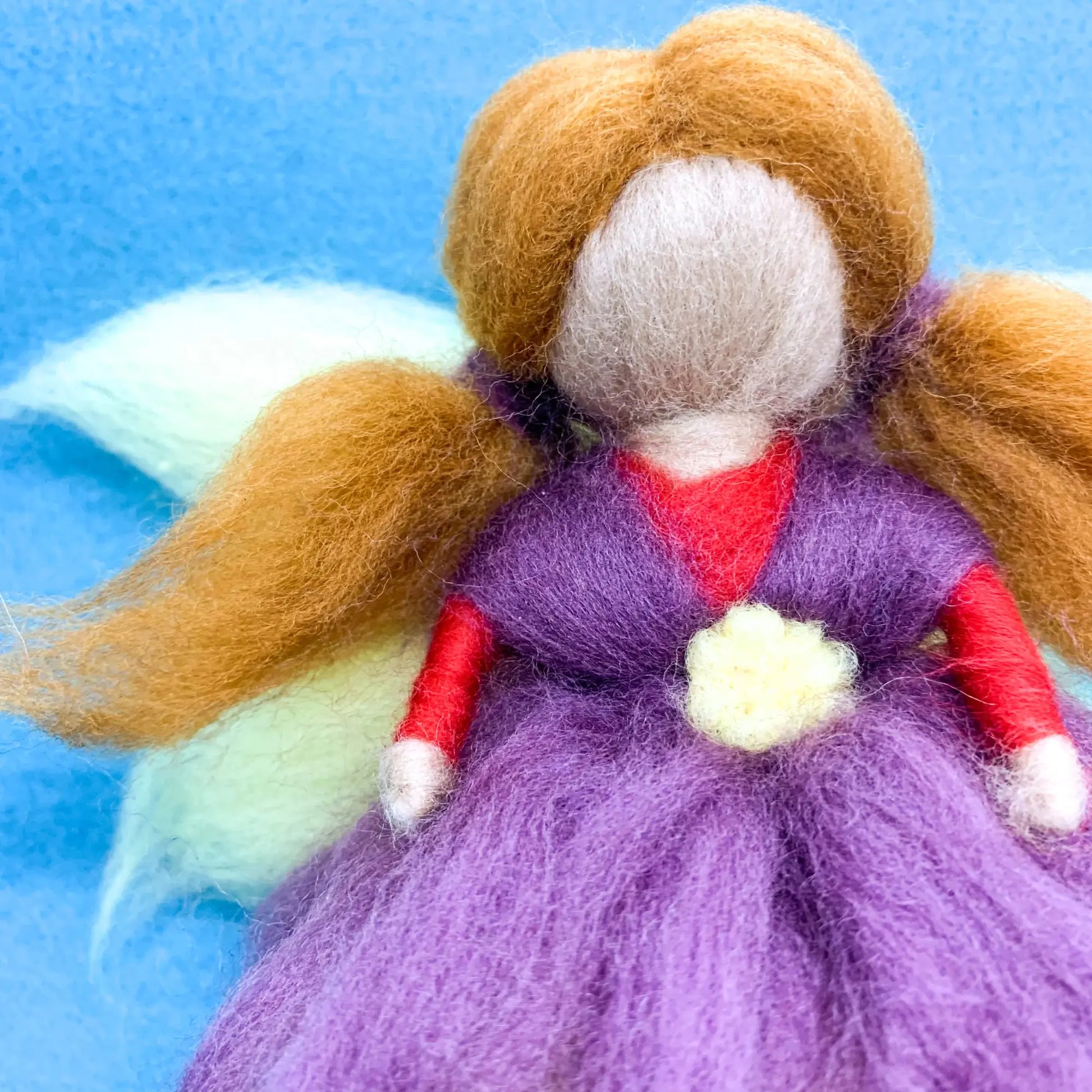 Felted fairy doll with brown hair, purple dress, and red shirt on a blue background