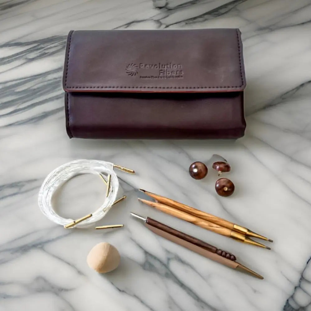Brown leather case with Revolution Fibers branding, accompanied by knitting needles and yarn on a marble surface.
