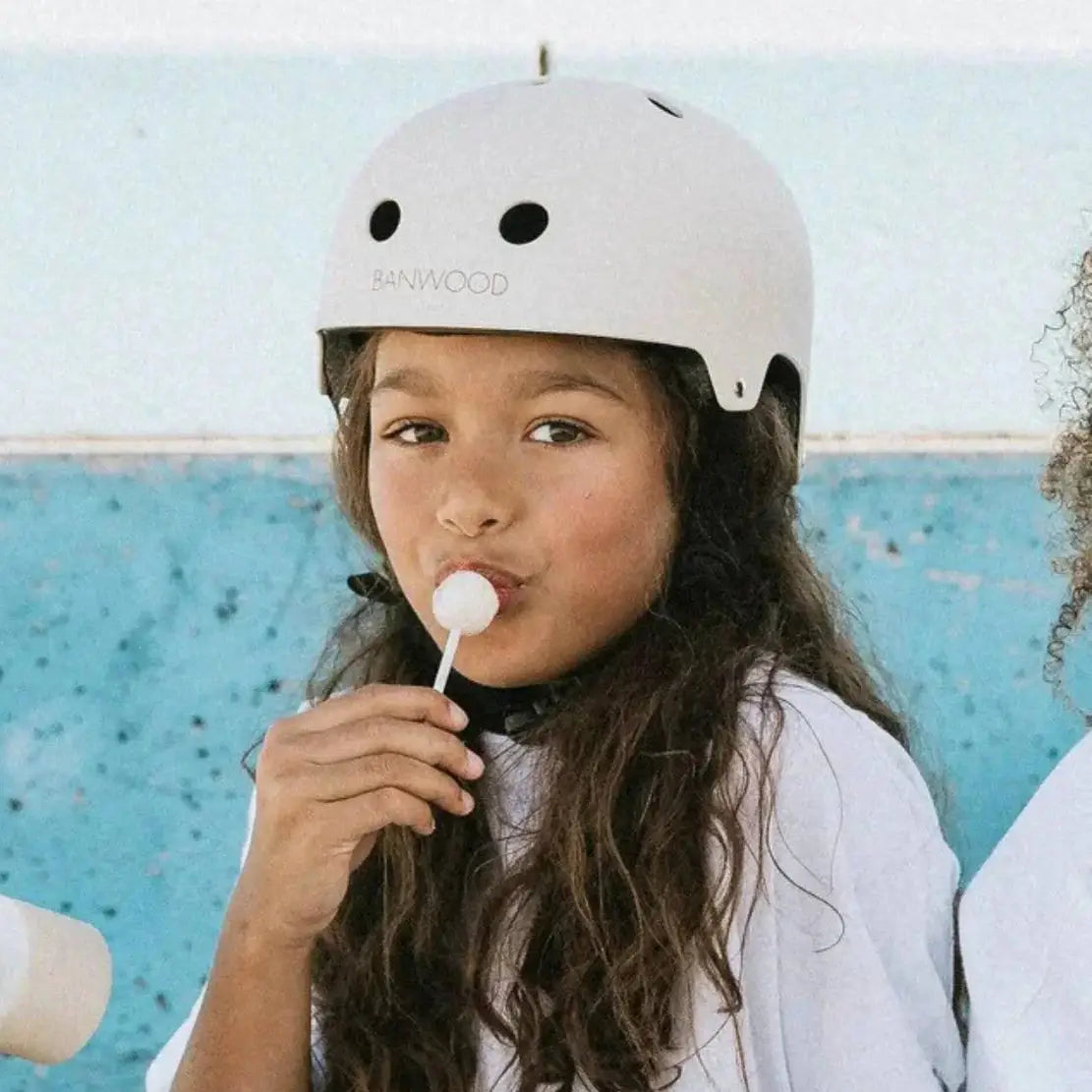 Child wearing a white helmet with 'Banwood' branding, eating a lollipop against a blue wall.