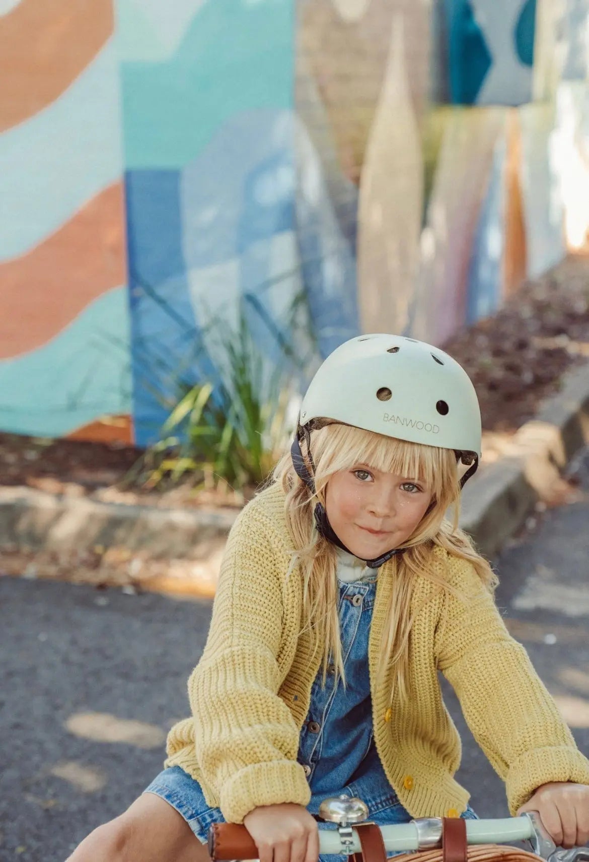 Child wearing a helmet and yellow sweater sitting on a bicycle in front of a colorful mural.