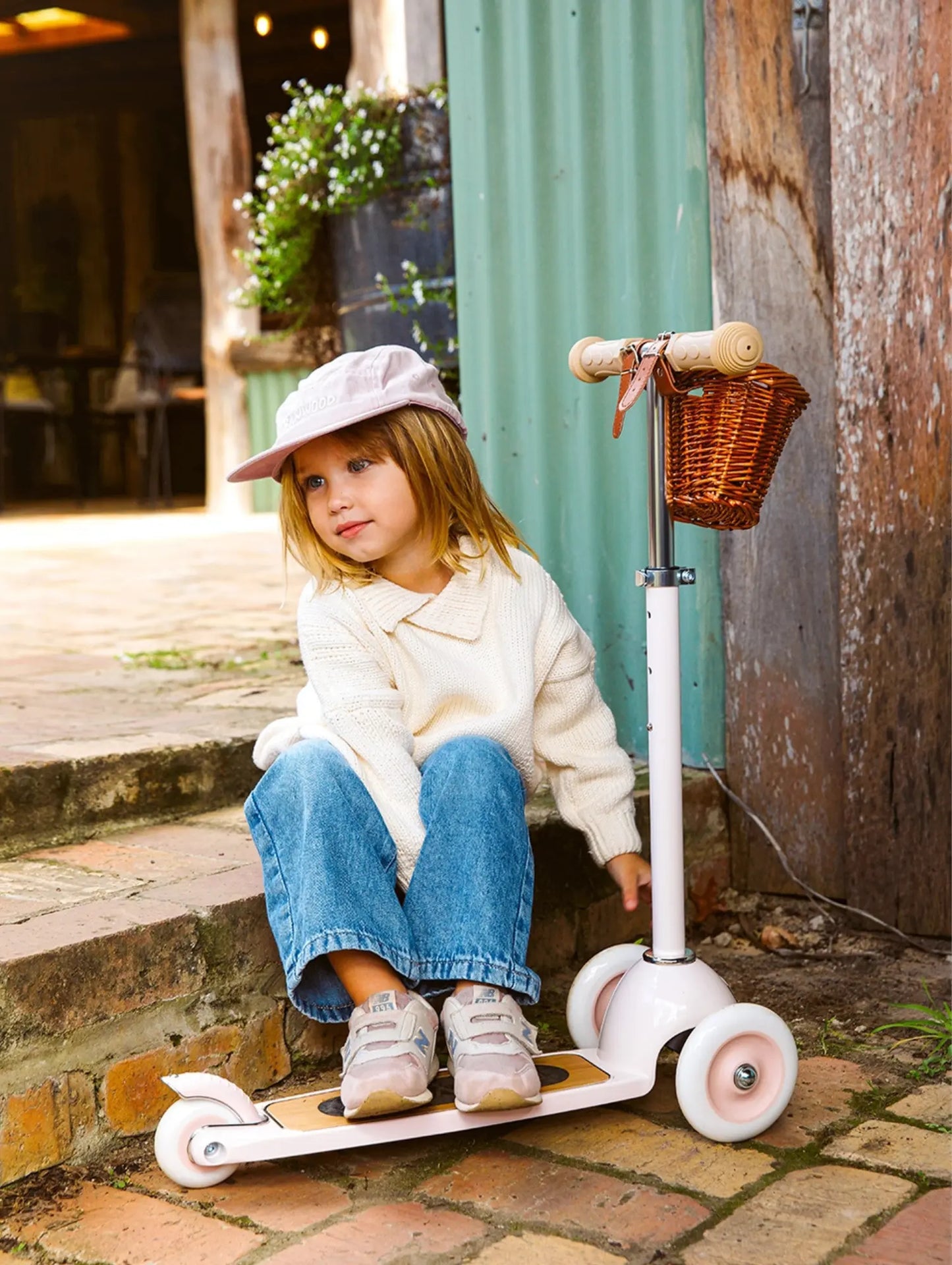 Child sitting on a scooter with a basket, wearing a white coat and pink hat, in an outdoor setting.