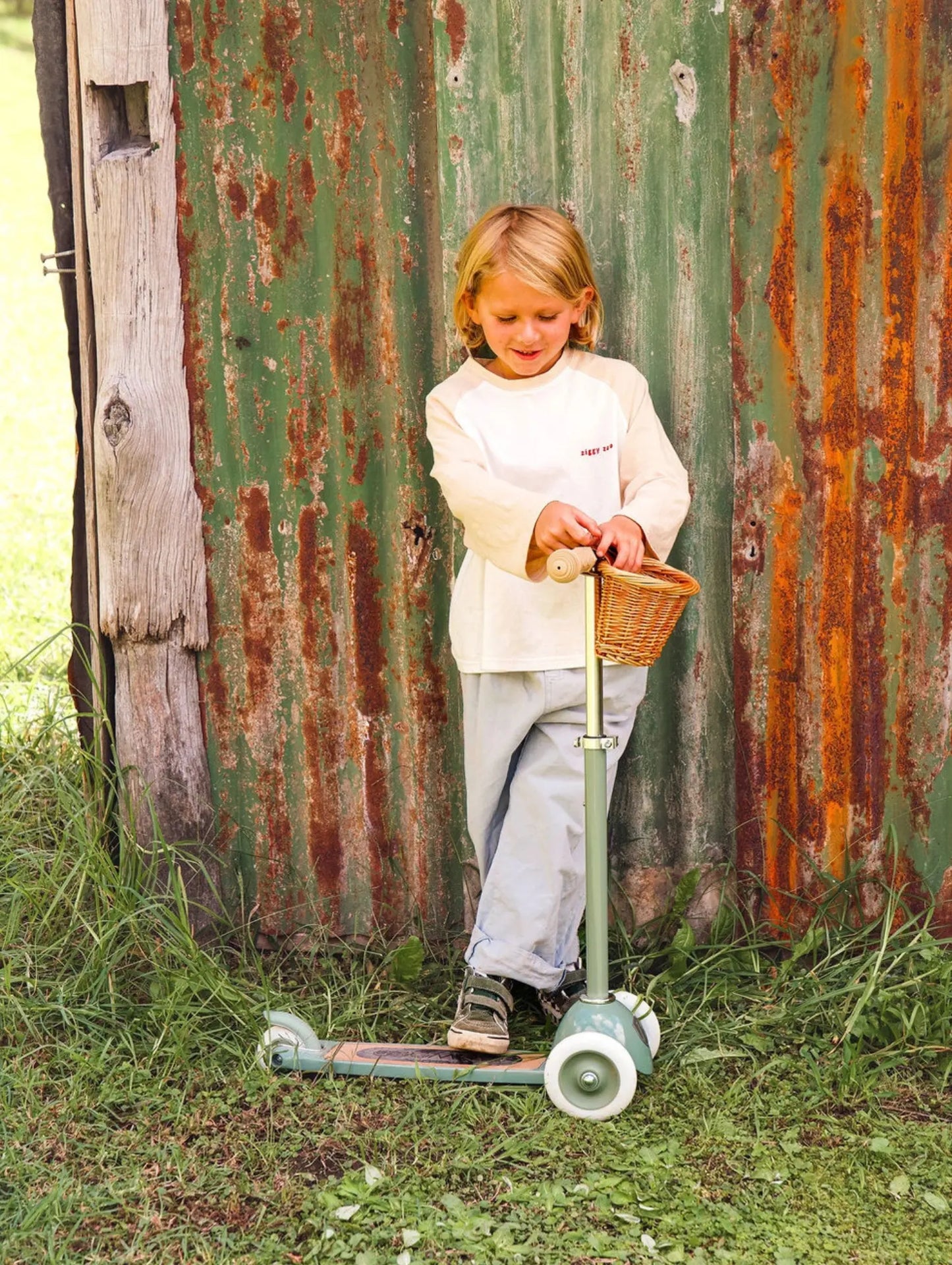 Child with a scooter and basket in front of a rusty metal wall