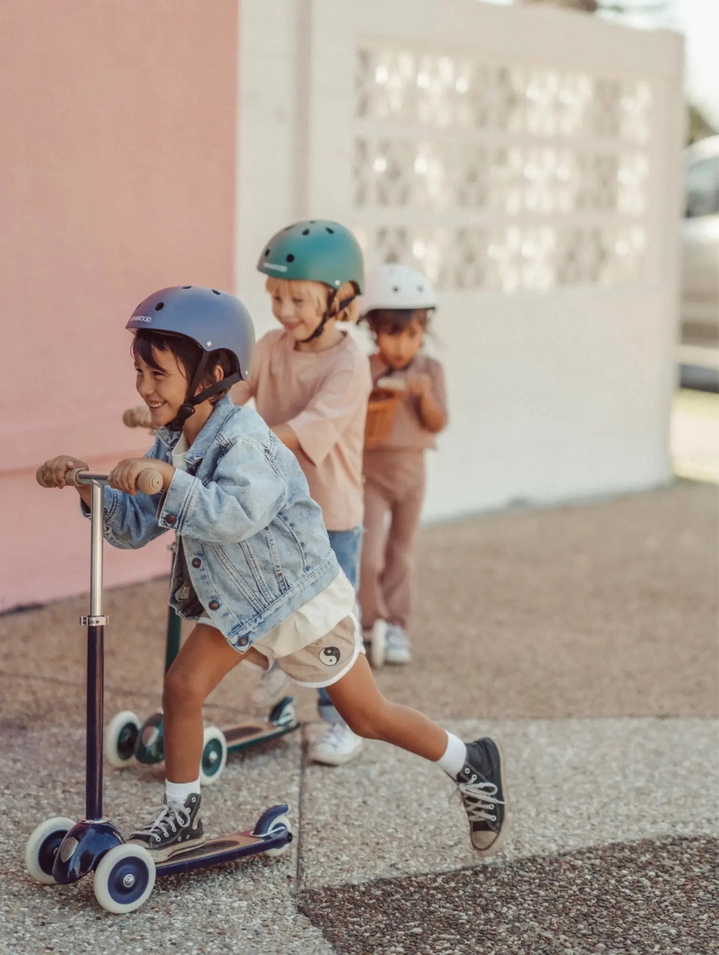Children riding scooters and skateboards with helmets on a sidewalk.