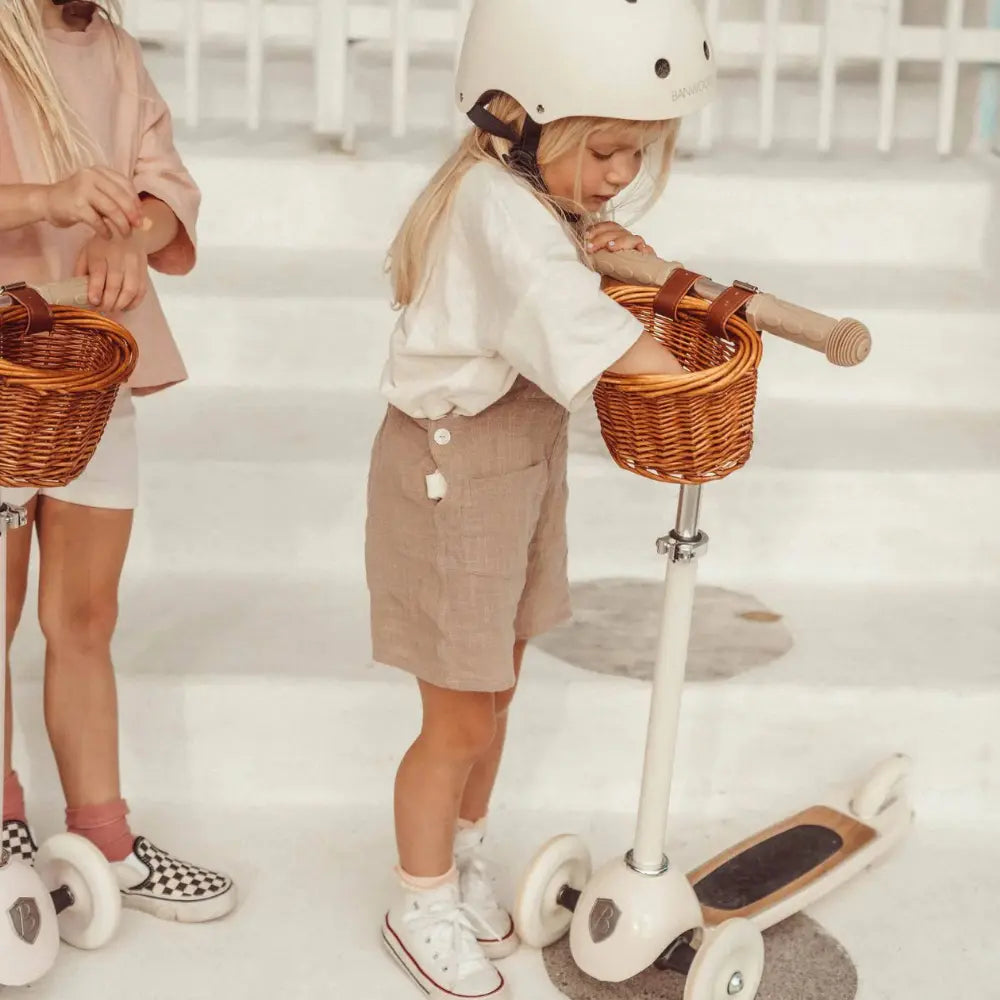 Two children on scooters with wicker baskets in a minimalistic setting