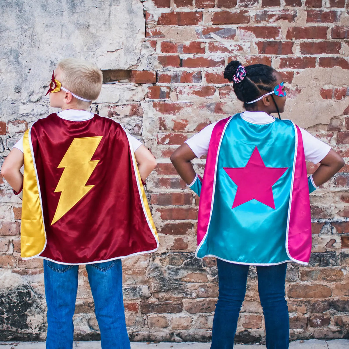 Two children wearing burgundy lightning bolt and teal star superhero cape sets during dress-up play.