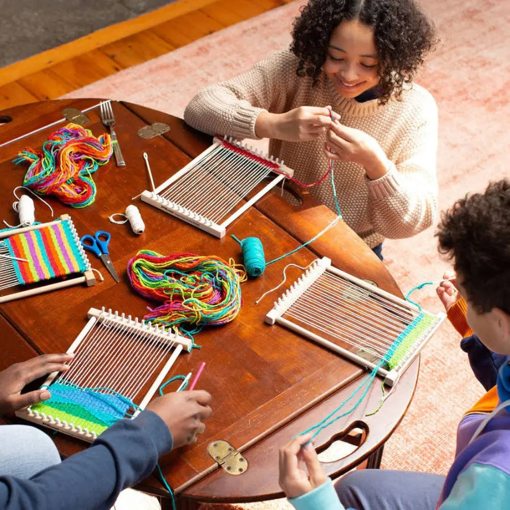Two people working on weaving projects with colorful yarn at a wooden table.