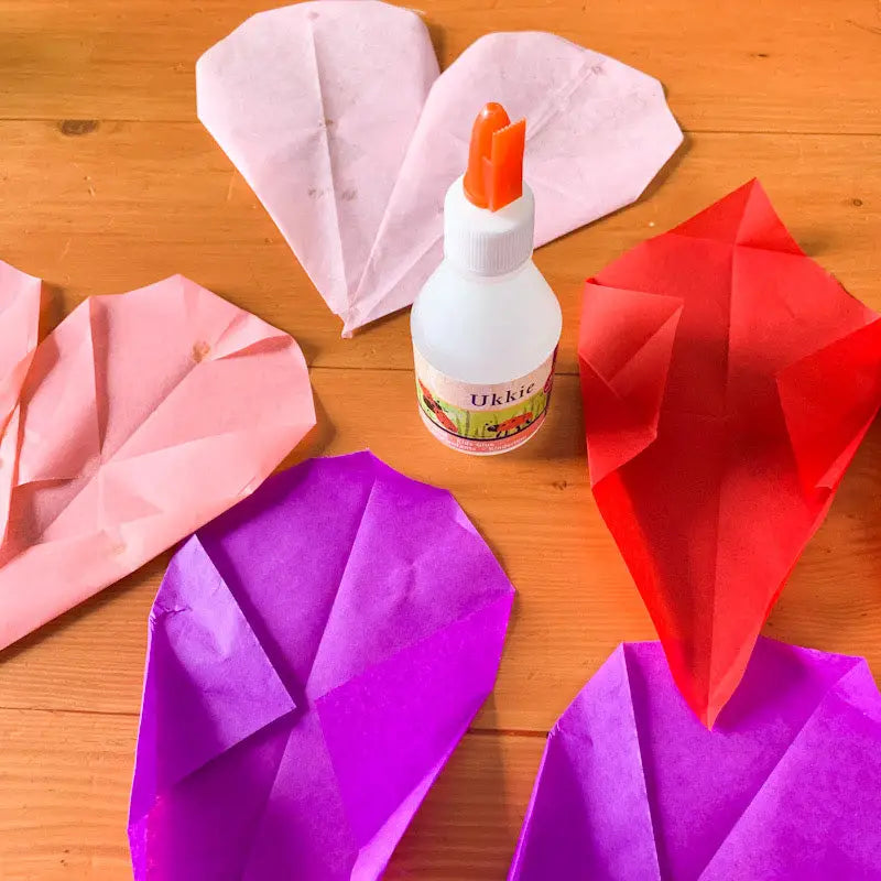 Valentine craft heart kit silk paper in red, purple, pink folded in different stages with a bottle of glue on a table