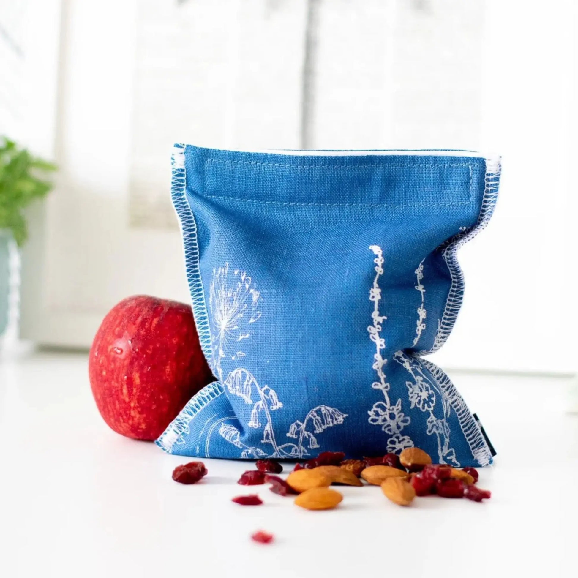 Blue fabric linen snack bag with white patterns next to an apple and snacks on a white surface