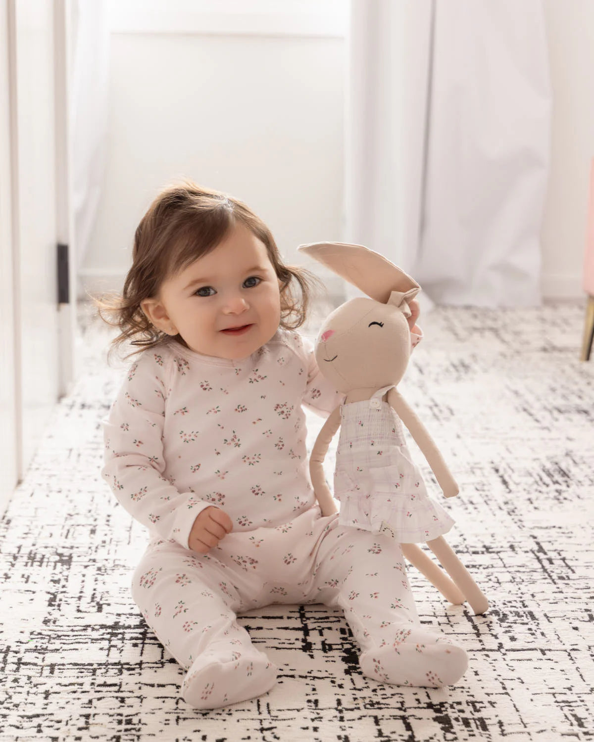 Baby in floral pajamas sitting on a patterned rug with a plush woodland bunny doll toy.