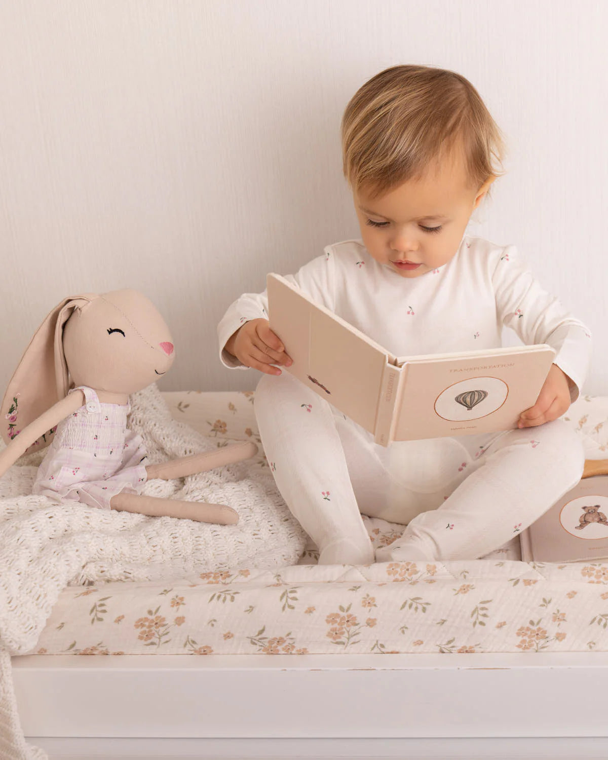 Child reading a book next to a plush bunny toy on a bed.