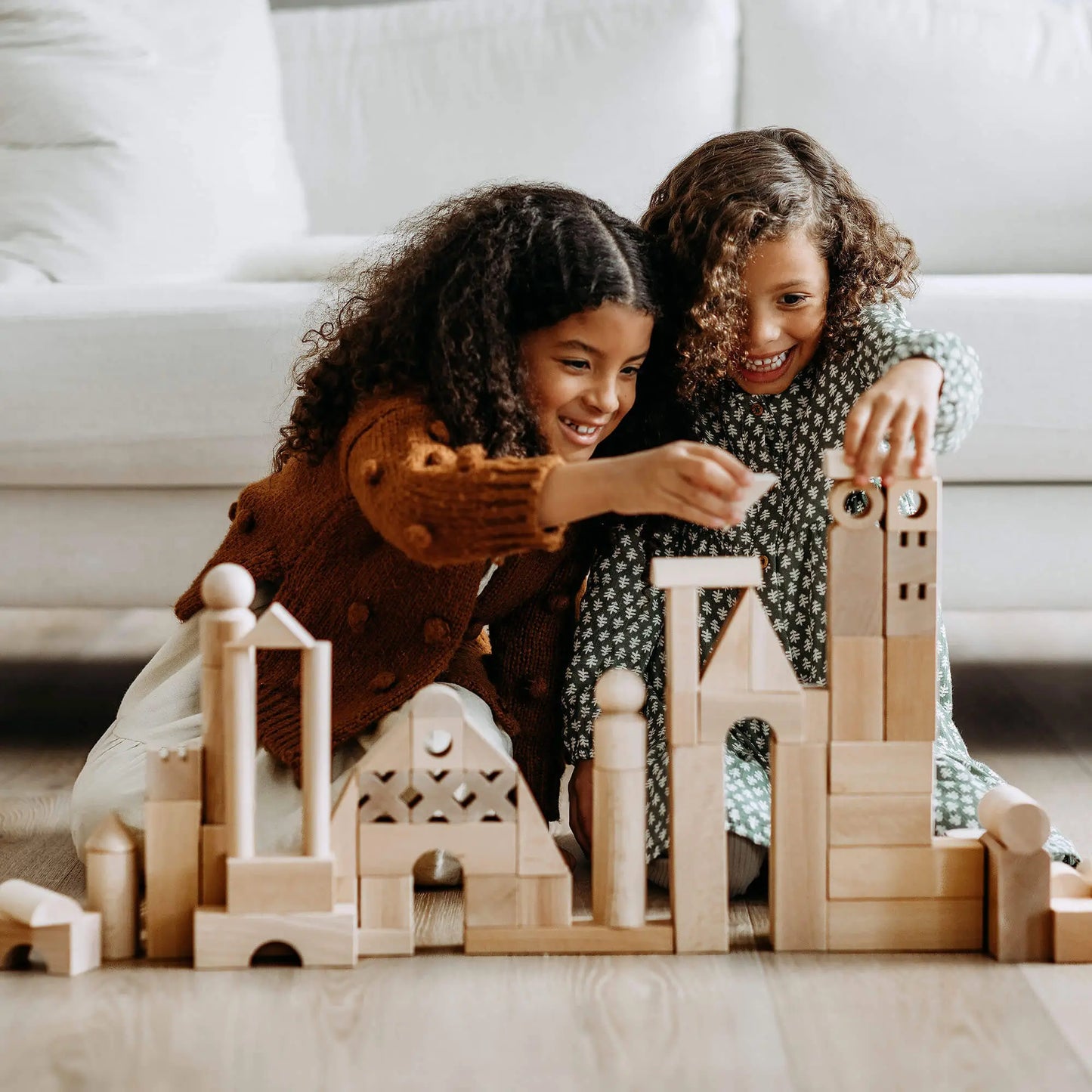 Two children playing with plain wooden building blocks on a light-colored floor.