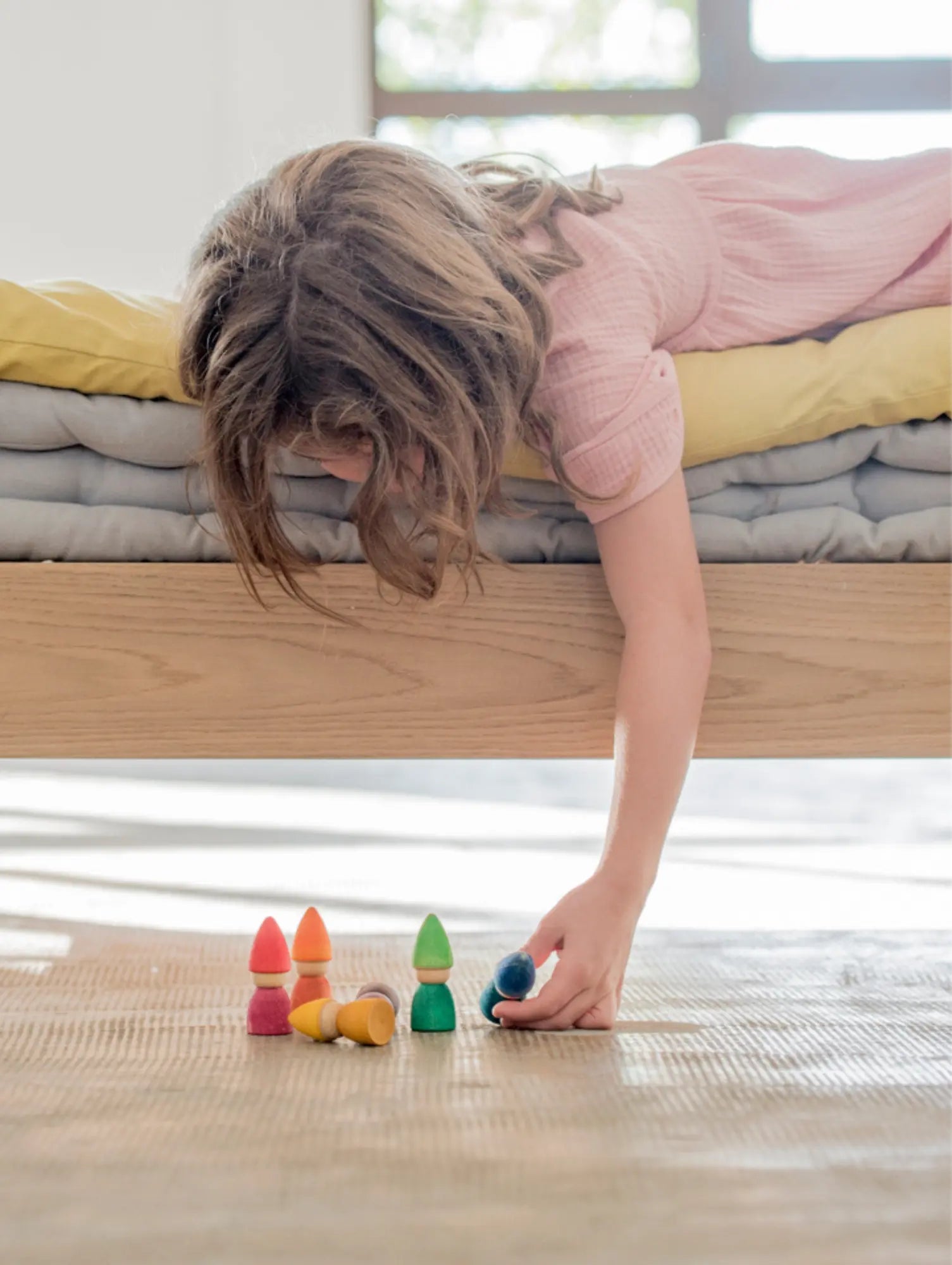 Child playing with colorful Grapat Wooden Tomtens on a wooden floor next to a bed.