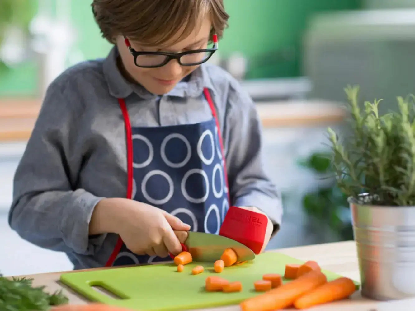 Child in a kitchen cutting carrots on a green cutting board with a red knife.