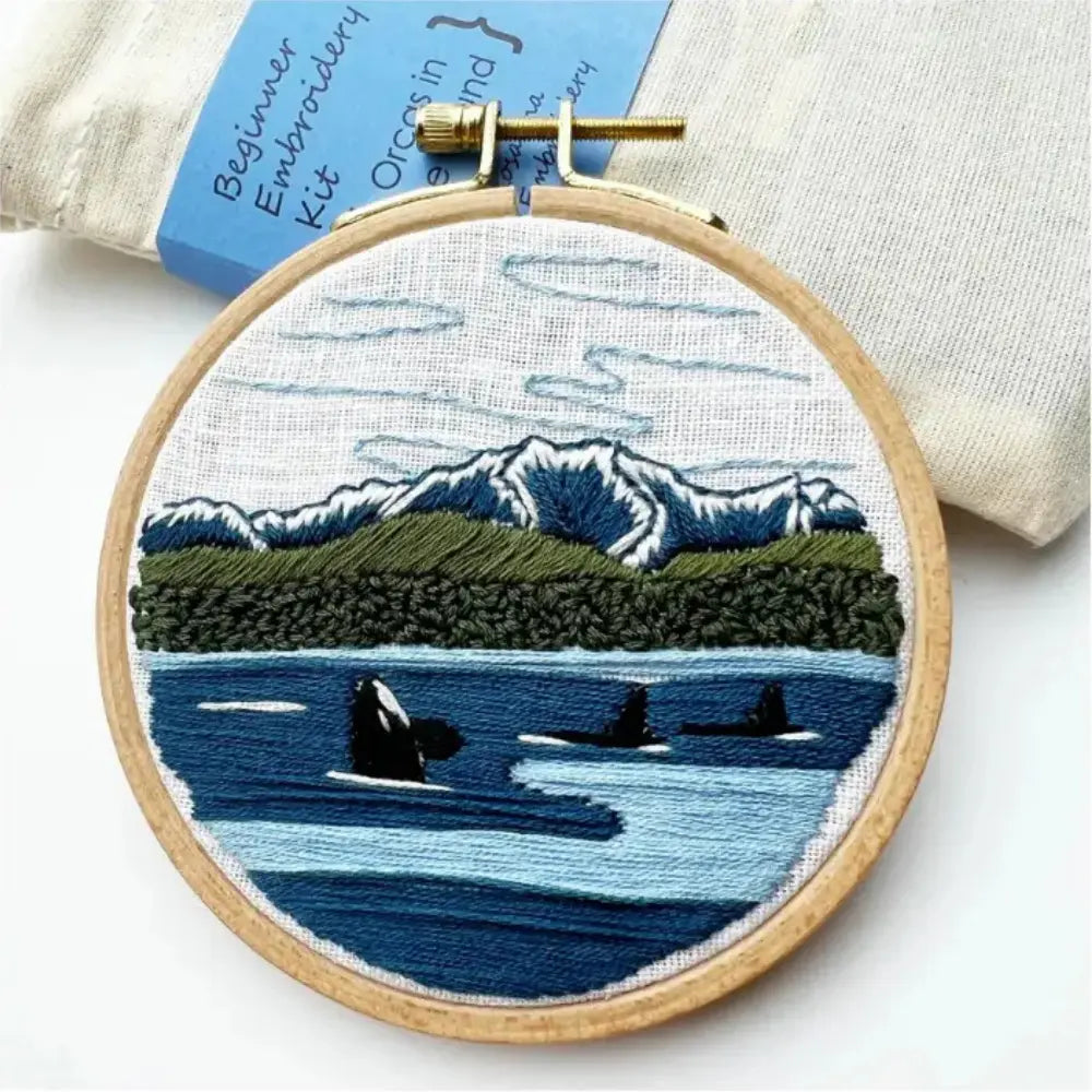 Embroidery hoop with a scenic design of mountains, trees, and water with orcas, on a white background.